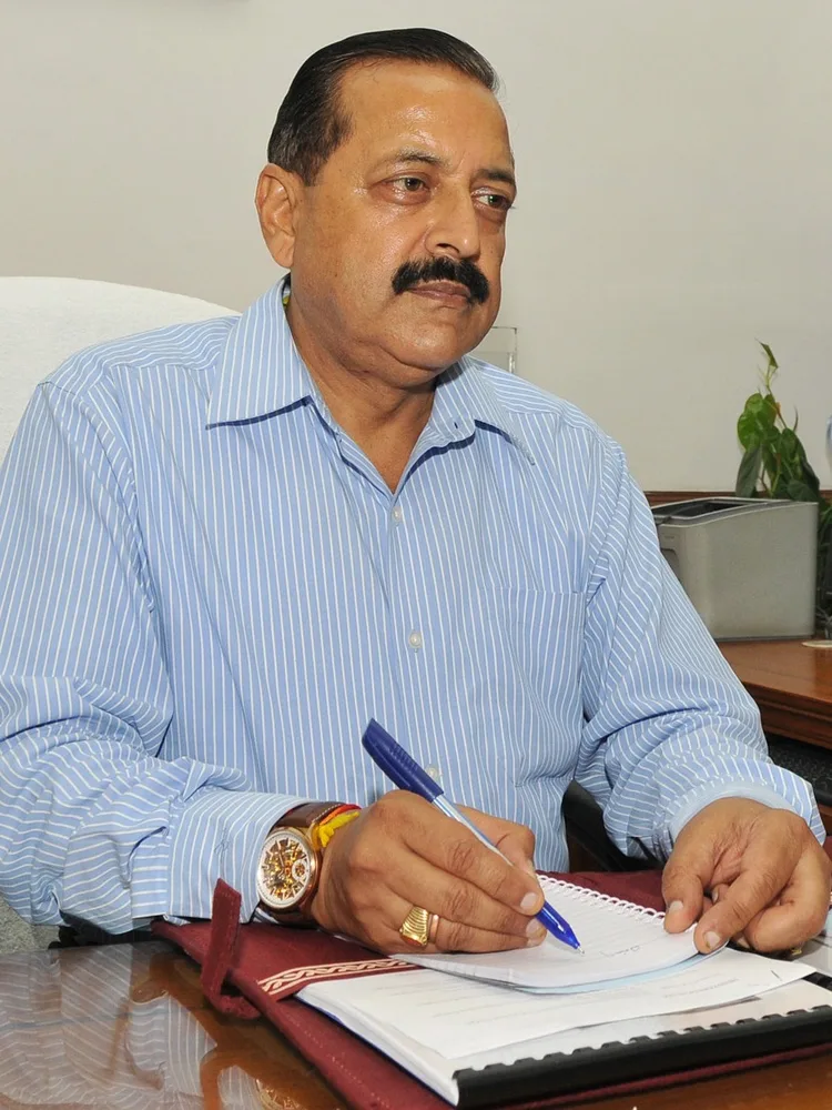 Dr. Jitendra Singh in a striped shirt seated at a desk, writing with a pen