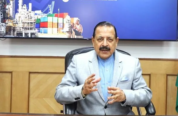Dr. Jitendra Singh in a grey jacket seated at a desk, gesturing, with a screen behind him