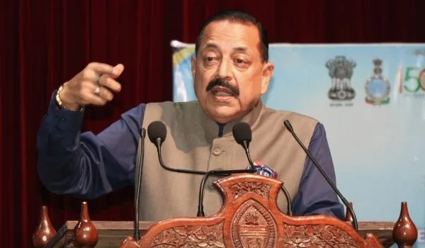 Dr. Jitendra Singh speaking at a carved wooden lectern with microphones, gesturing with his right hand