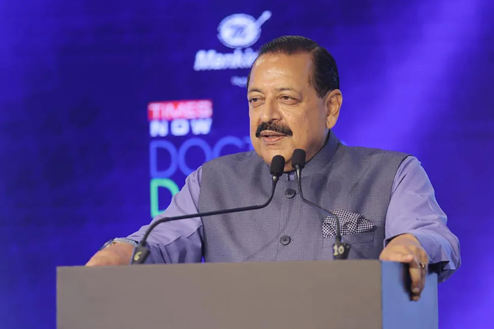 Dr. Jitendra Singh speaking at a lectern with microphones against a blue-lit stage backdrop