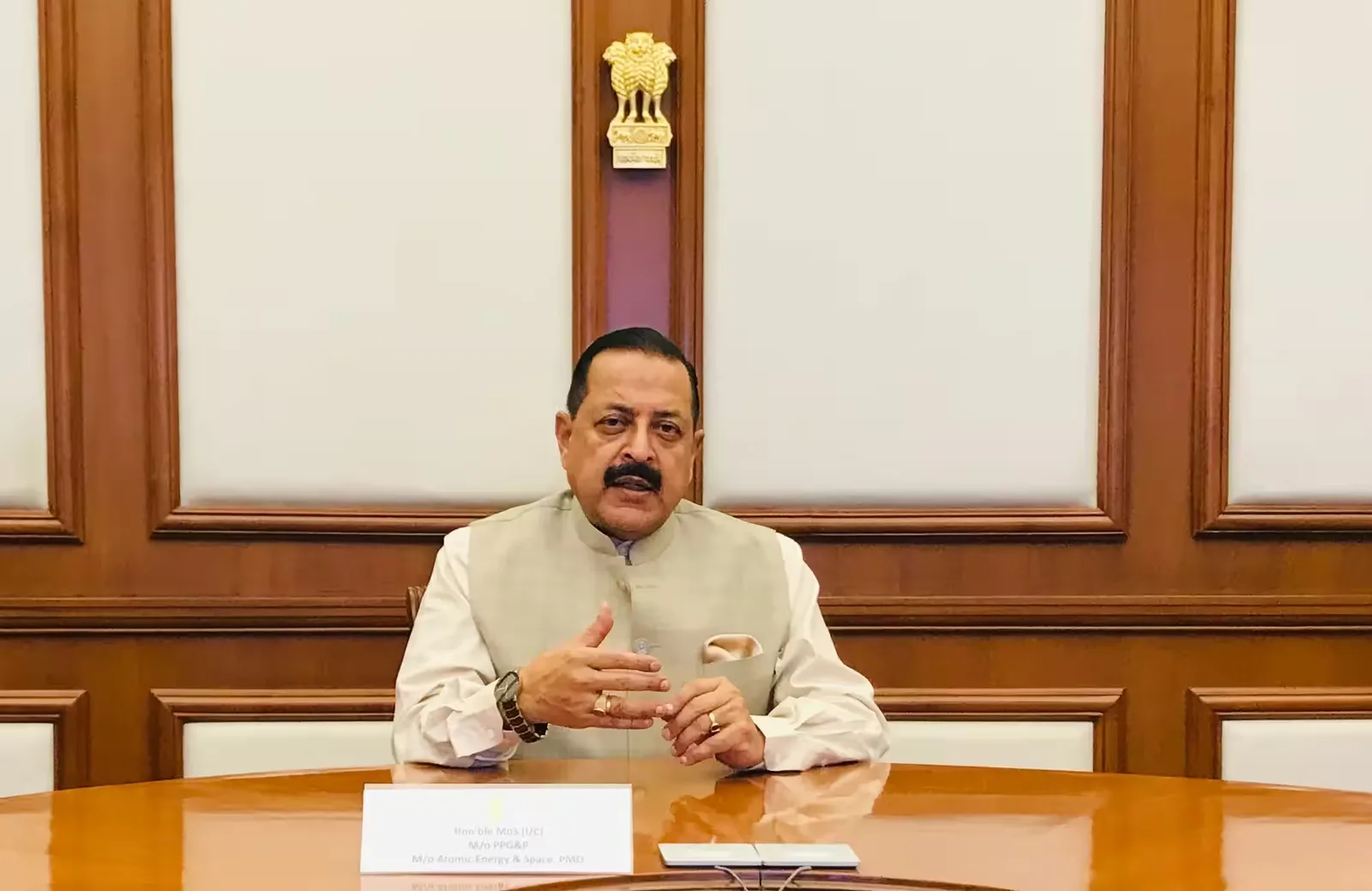 Dr. Jitendra Singh seated at a polished table in a wood-panelled room beneath the national emblem, speaking
