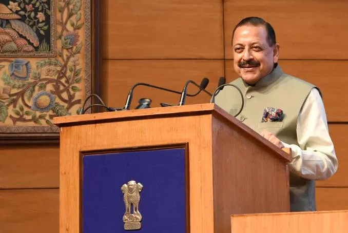 Dr. Jitendra Singh smiling while speaking at a wooden lectern with microphones, in front of a textile artwork